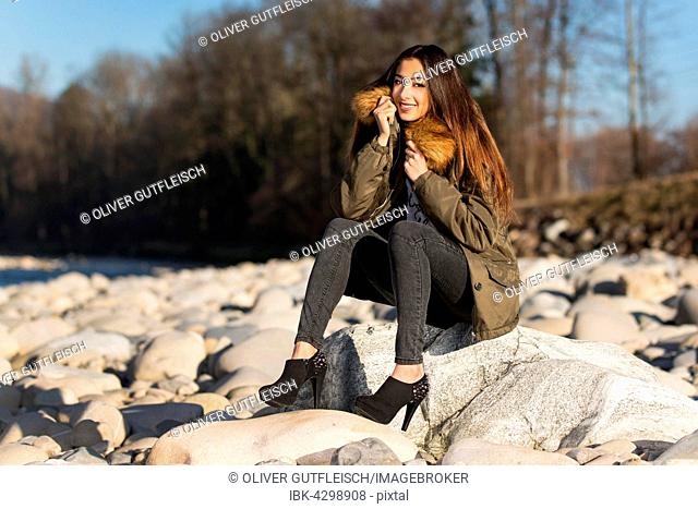 Young woman with long brunette hair sits on stone in dry river in winter, fashion, look into the camera