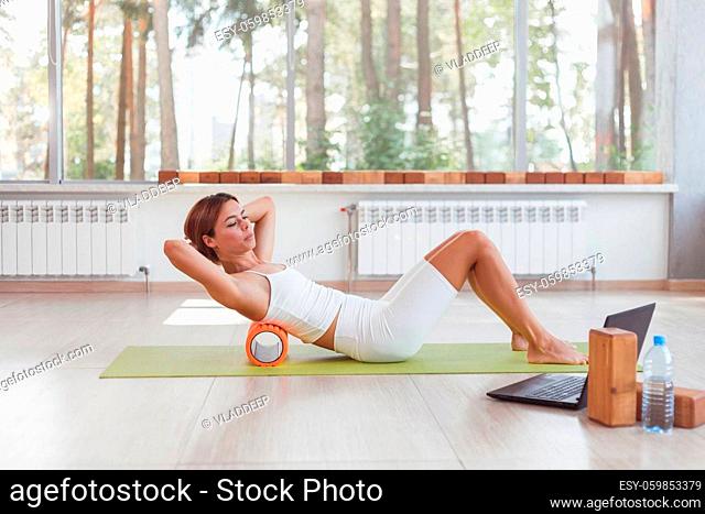 Sporty woman doing online exercises with foam roller under the back. Workout online with laptop. Instructor online