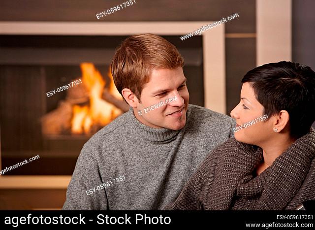 Young couple hugging on sofa in front of fireplace at home, looking at each other, smiling