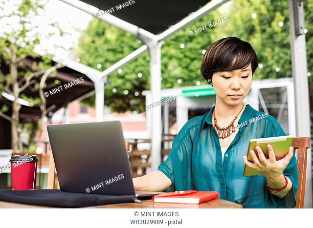 Woman with black hair wearing green shirt sitting in front of laptop at table in a street cafe, holding digital tablet