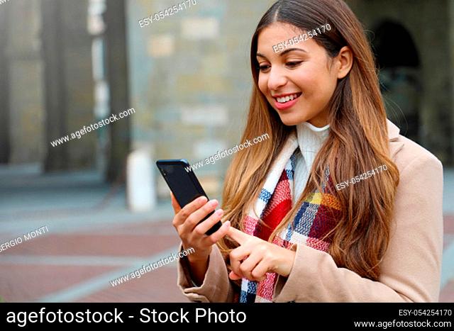 Portrait of beautiful young woman with coat and scarf in city typing on her phone