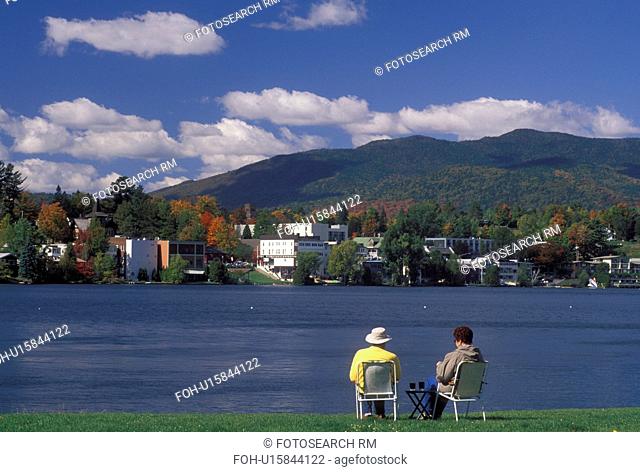 Lake Placid, NY, New York, The Adirondacks, Couple sitting on the lakefront of Mirror Lake looking at the resort town of Lake Placid in the autumn