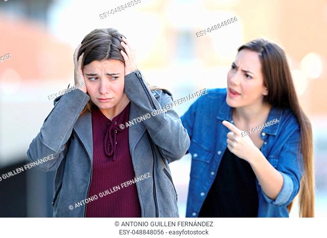 Angry girl accusing her sad friend who is covering ears walking in the street