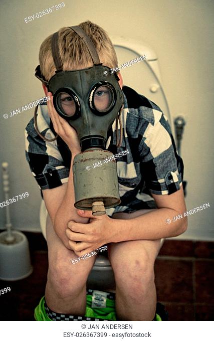 Single blond boy in gas mask sitting on toilet in bathroom with pants down and hands on face