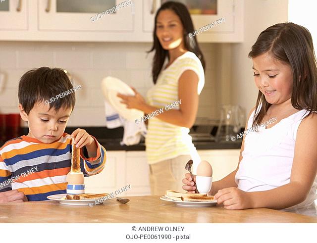 Two young kids in kitchen eating eggs and toast with woman in background smiling