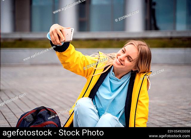 Beautiful girl takes selfie on smartphone and sits on skateboard, against the background of the street