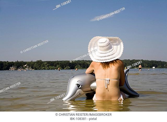 Young woman with an inflatable dolphin and a large white sun hat looks into the distance, view from the back at the bathing beach Wannsee, Berlin, Germany