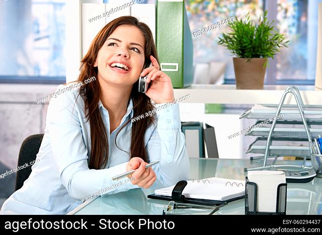 Girl sitting in office speaking on mobile phone, holding pen, laughing
