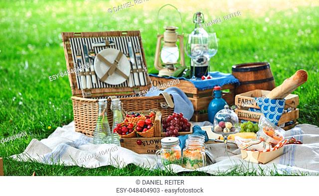 Vintage style picnic hamper with lunch in a park spread out on a rug on the grass with cheese, fresh fruit, bonbons, pickles bread and red wine