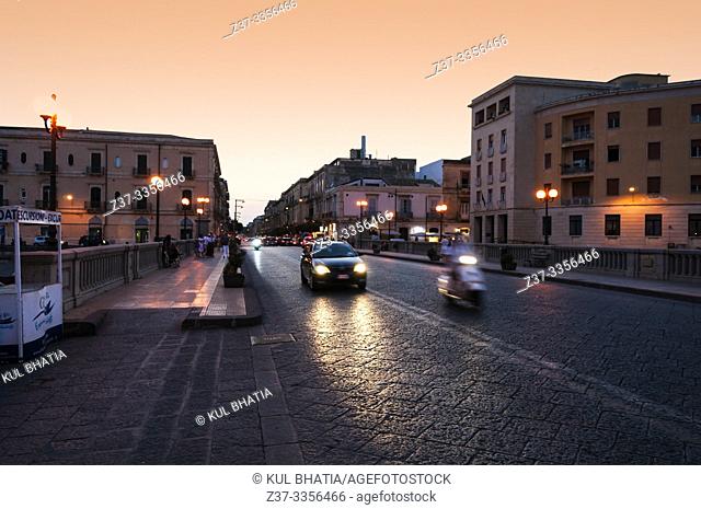 Vittorio Emmanuel bridge, connecting Ortigia and Siracusa at dusk. Dark stones in the pavement glow in the headlights of cars and scooters on a warm summer...