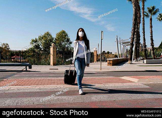 Woman with luggage crossing street against blue sky on sunny day during pandemic