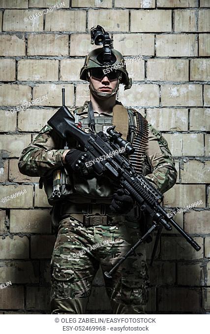 US Army Ranger with machinegun and night vision goggles standing near the wall. Front view portrait