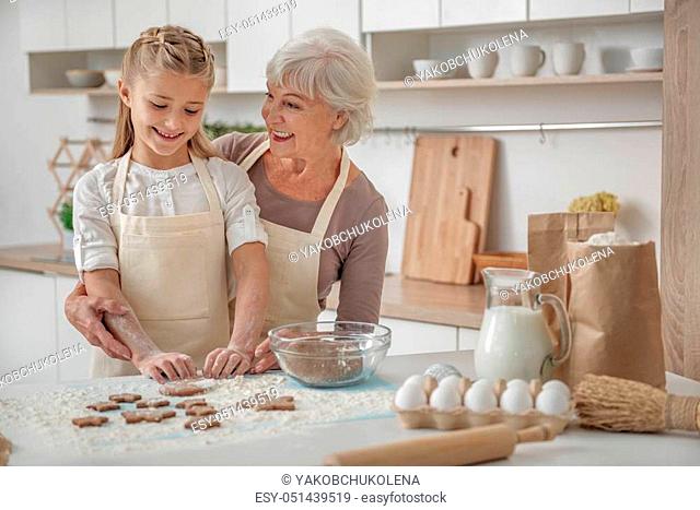 Well done. Happy senior woman is baking cookies with her lovely granddaughter. She is embracing the girl with proud and smiling