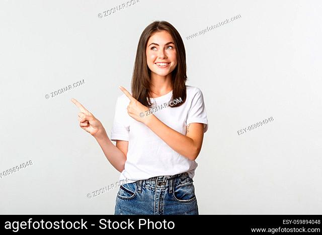 Dreamy beautiful girl smiling, pointing fingers upper left corner and looking pleased at logo