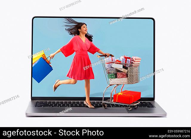 A young woman carrying colorful shoppingbags moving a cart full of gift boxes within the graphics of laptop screen