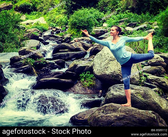 Woman doing yoga asana Natarajasana, Lord of the dance pose outdoors at waterfall in Himalayas. Vintage retro effect filtered hipster style image