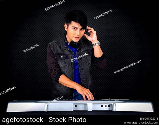 Young asian man with headphones playing an electric keyboard in front of black soundproofing wall. Musician producing music in professional recording studio