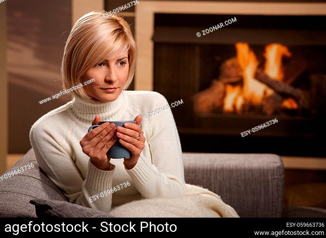 Woman sitting on sofa at home drinking hot tea, looking away