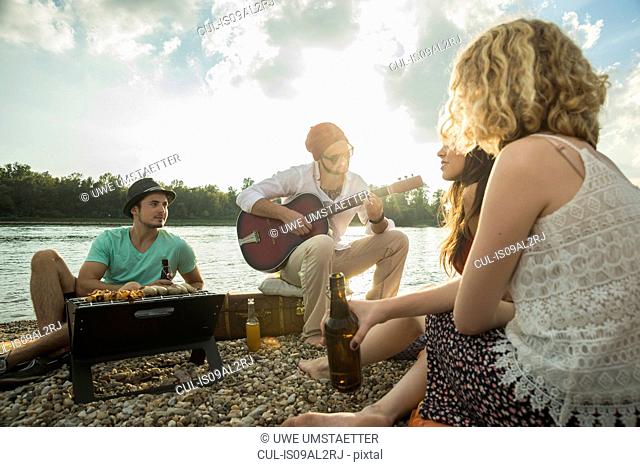 Young man sitting by lake with friends playing guitar