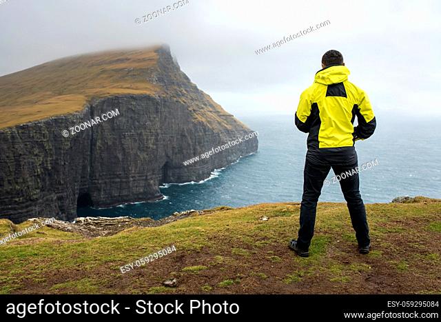 Guy in a yellow jacket stands on the cliff on the seaside on the foggy horizon background on Faroe Islands. Cliff covered with grass