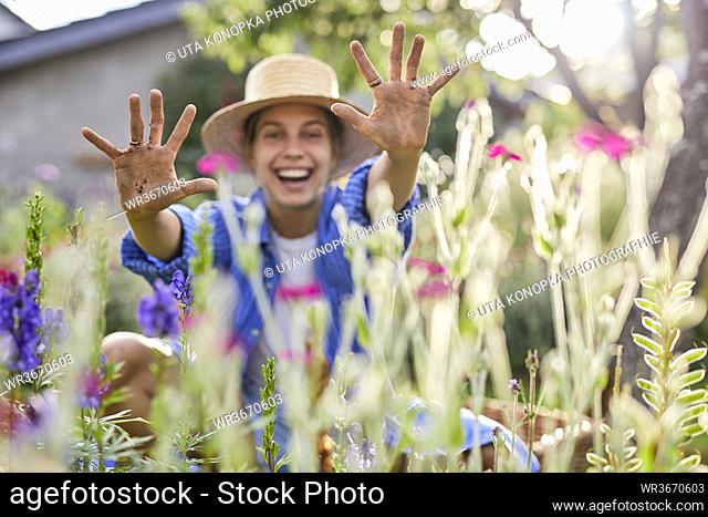 Cheerful woman wearing hat showing dirty hands while sitting amidst plants in garden