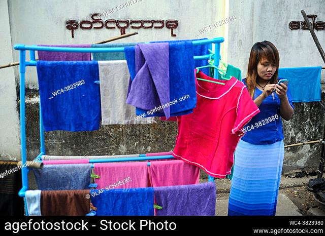 Yangon, Myanmar, Asia - A young woman stands next to a laundry rack with colourful towels and stares at her smartphone