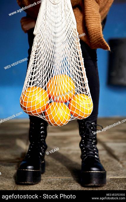 Young woman holding oranges in reusable bag while standing on footpath