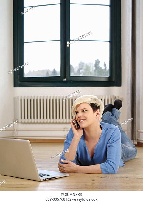 Photo of a beautiful young female lying on the floor working on a laptop and talking on a smartphone
