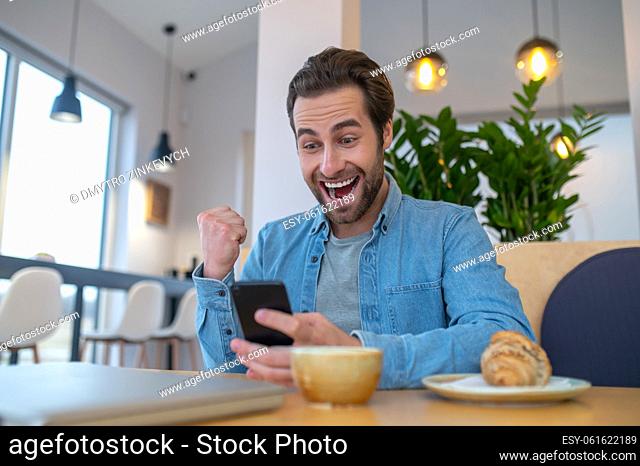 Joyful moment. Enthusiastic young bearded man with open mouth gesturing with fist looking at smartphone sitting at table in cafe