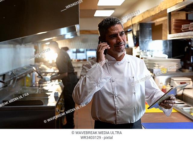 Front view close up of a middle aged Caucasian male chef on the phone and holding a tablet computer in a restaurant kitchen