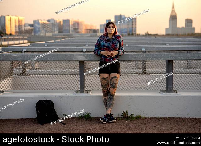Fashionable woman day dreaming while leaning on railing at rooftop during sunset