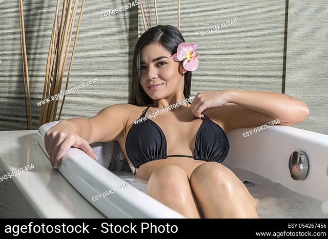 Flirty young brunette in black bikini lying in healing bath in massage center smiling at camera