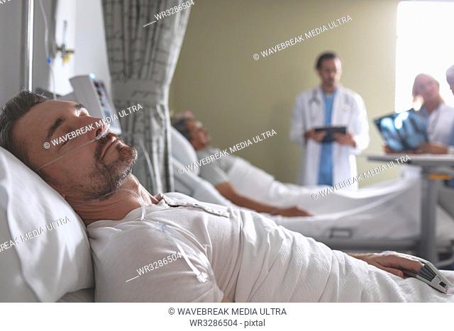 Side view of Caucasian male patient sleeping on bed in the ward at hospital. In the background diverse doctors discussing x-ray with mixed-race male patient