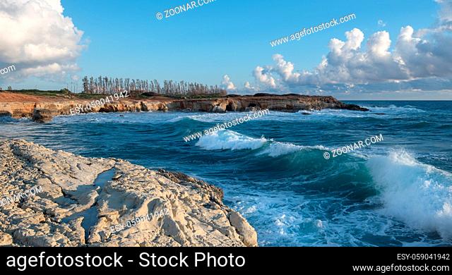 Sea waves splashing on the rocks of a rocky beach against blue cloudy sky. Paphos Cyprus