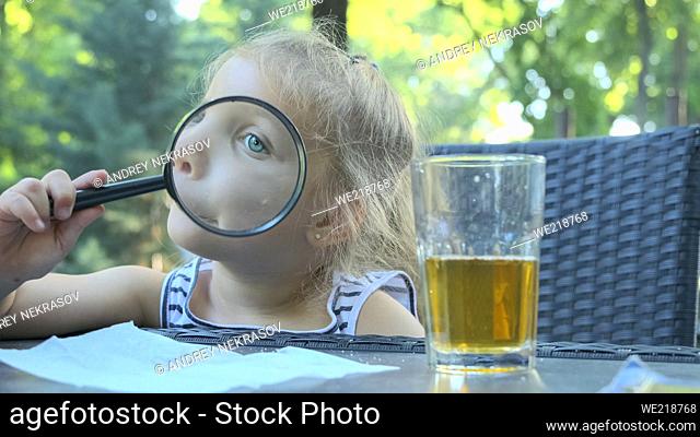 Little girl looks into the lens around. Close-up of blonde girl studying the world around her looking at it through magnifying glass while sitting in street...