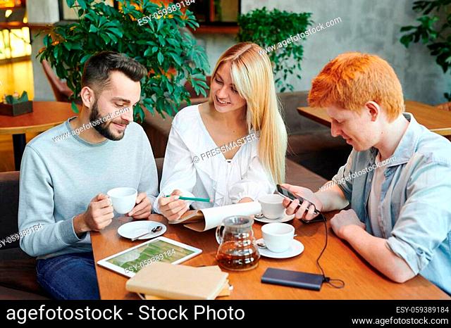 Pretty blonde girl showing her boyfriend notes in notepad while both reading them and preparing for seminar by cup of tea in cafe