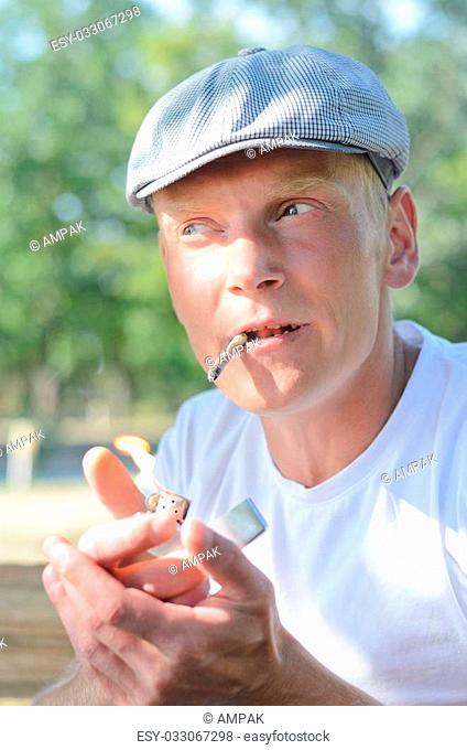 Young man in a cap about to light a self rolled cigarette or joint which is dangling from his mouth sitting outdoors in the shade of a tree
