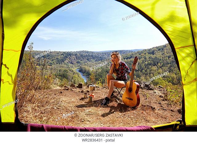 Young blonde woman tourists with guitar in camp on cliff over river and forest landscape