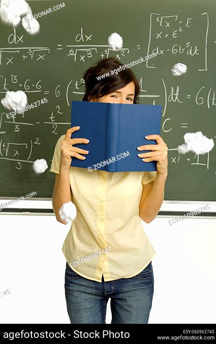 Teenage girl standing in classroom, in front of blackboard. Hiding behind book. Someone throwing pieces of paper into her. Front view