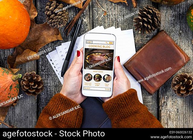 Female holding smartphone with online cooking blog on the screen in the kitchen with notepad, pumpkins and leaves on wooden table. Autumn composition