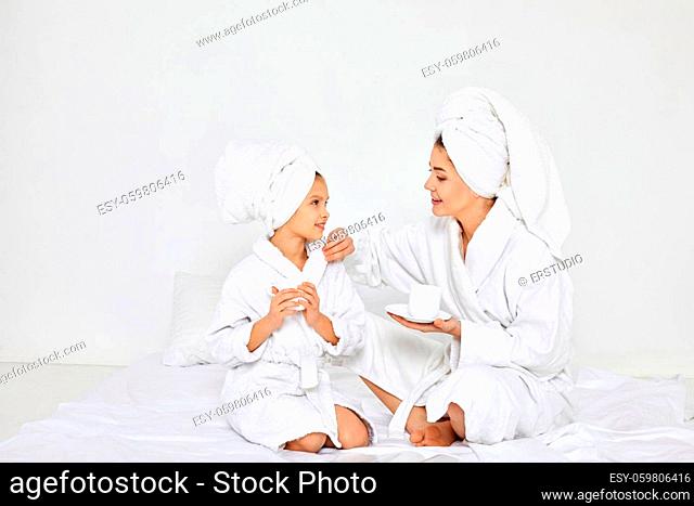 happy mother and daughter in white bathrobes and towels on heads drinking tea in bedroom