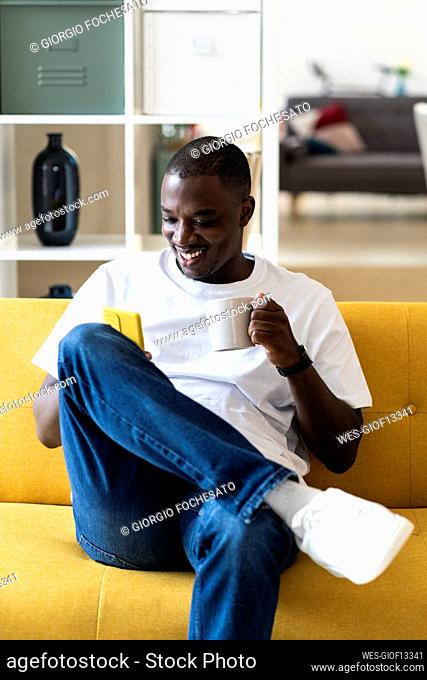 Man holding mug using smart phone while sitting on sofa at home