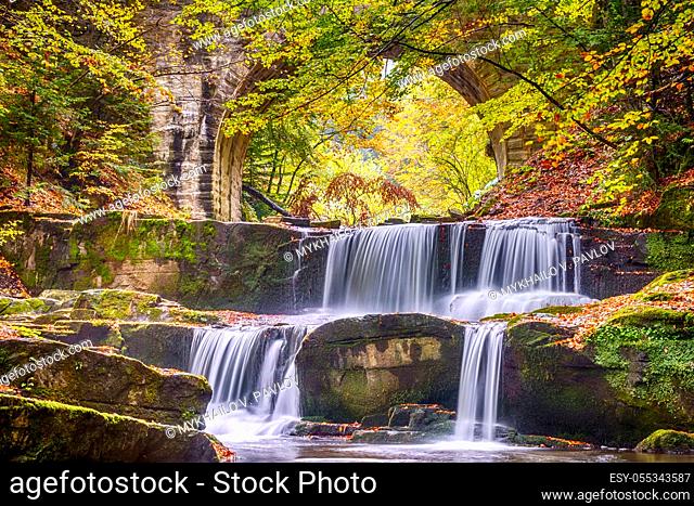 Autumn day in the sunny forest. Old stone bridge. Small river and several natural waterfalls