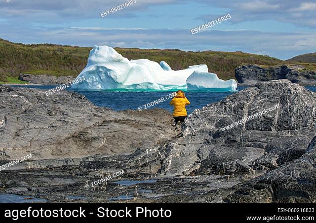 A female tourist in a bright yellow coat photographs an iceberg near Noddy Bay, Newfoundland