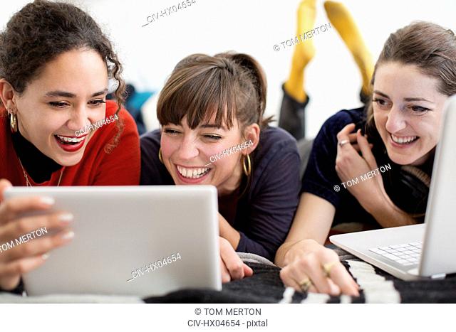 Laughing young women friends hanging out, enjoying digital tablet and laptop on bed