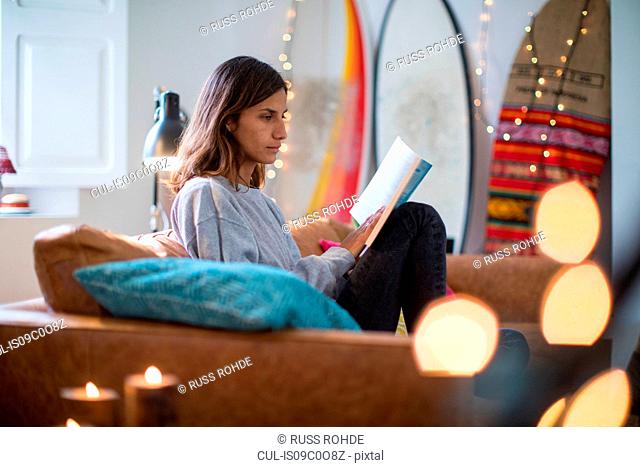 Young woman sitting on living room sofa reading a book