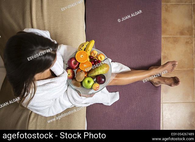 From above shot of woman in white bathrobe sitting on bed with plate of colorful fruit