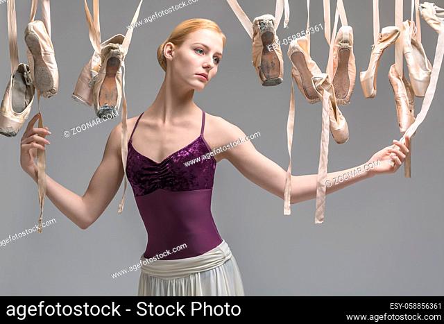 Beautiful ballerina in a violet top and a cream skirt stands on the gray background in the studio. Around her there are many hanging beige pointe shoes