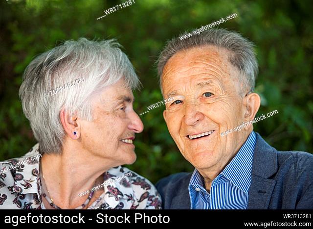 Head-and-shoulder portrait of an 80 year old pensioner couple against blurred green background. Woman looks at man. Man looks to the camera