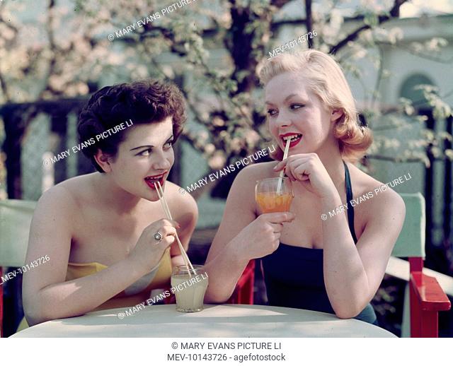 Two girls in bathing costumes sip soft drinks through straws as they sit at an outside table enjoying the sunshine